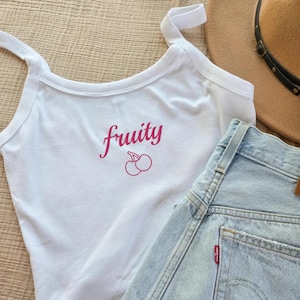 Puede incluir: Camiseta sin mangas blanca con la palabra "Fruity" en escritura rosa y un gráfico de cereza. Pantalones cortos vaqueros azul claro y un sombrero marrón también están en la imagen. Los artículos están dispuestos sobre una superficie de color neutro.