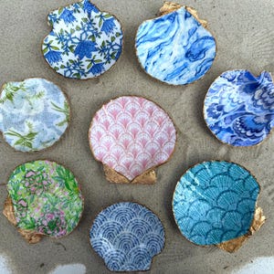 May include: A collection of eight decorative scallop shells, each featuring a unique pattern including blue floral, marbled blue, pink fan, and teal scale designs. Each shell has a gold-painted edge, arranged in a circular formation.