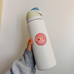 May include: White reusable water bottle with a blue and beige lid. The bottle has a sticker on it that says "cherry Coke" in red and white letters.