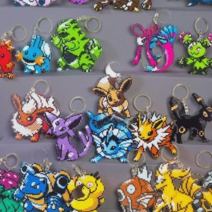 May include: A collection of colorful pixel art keychains featuring various Pokémon characters. Each keychain has a metal ring for attachment. The keychains are arranged on tiered gray platforms, showcasing a variety of designs and colors.