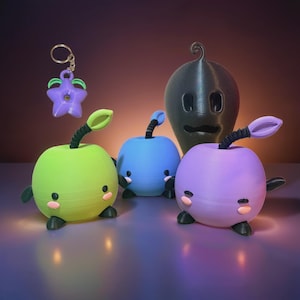 May include: A collection of whimsical, colorful 3D-printed figures. Includes a green, blue, and purple apple-shaped character with black stems and tiny feet. A black, gourd-shaped figure and a purple star-shaped keychain are also present.
