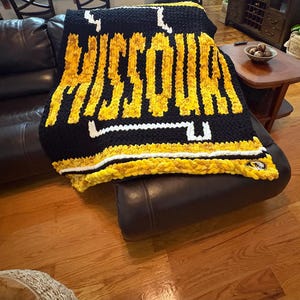 May include: A chunky knit blanket featuring the word MISSOURI in yellow yarn against a black background. The design includes white accents, a horizontal white stripe, and a small circular logo patch in the bottom right corner of the blanket.