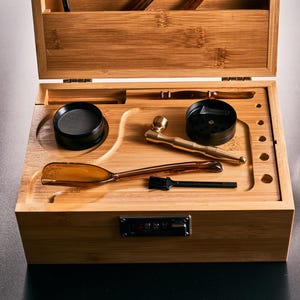 May include: A bamboo stash box with various accessories. The box includes a grinder, a pipe, a scoop, and two small glass jars with wooden lids. The box is open, displaying the contents.
