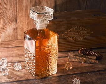 May include: A clear glass decanter with a square base and a diamond pattern. The decanter is filled with a golden liquid and has a personalized inscription that reads "Lord Will Conquer". The decanter is sitting on a wooden surface next to a wooden box with the inscription "Pendleton".