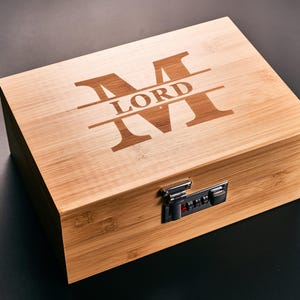 May include: A rectangular wooden box with a combination lock. The box is made of light-colored wood and features the word "LORD" and the letter "M" engraved on the lid. The box is closed and sits on a dark surface.