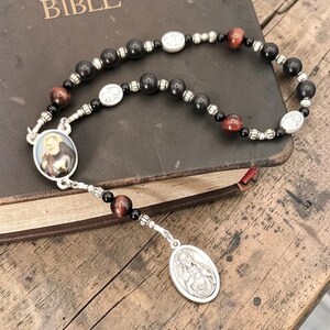 May include: A rosary with black and reddish-brown beads, silver-toned spacers, and oval medallions. The rosary is resting on a closed, brown book with the word "BIBLE" visible on the cover. The beads are made of a dark material.
