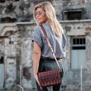 May include: A burgundy leather crossbody bag with a long, adjustable strap. The bag features a decorative fringe design and is secured with a buckle. The bag is worn over the shoulder.