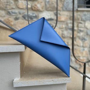 May include: A vibrant blue clutch bag with a unique triangular shape. The bag is made of smooth material and features a folded flap closure. The bag is resting on a set of stone steps.