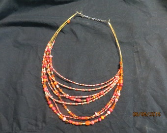 May include: Multi-strand necklace with red, orange, and gold beads. The necklace is made of multiple strands of beads, with a gold chain closure.