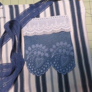 May include: A blue and white striped tote bag with a blue denim pocket. The pocket has white lace trim and a scalloped edge. The bag has a blue crocheted strap.