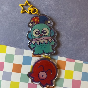 May include: Two acrylic keychains featuring cartoon characters. One is a teal clown-like figure with a colorful hat and a star-shaped clasp. The other is a red octopus with a single eye. Both have a glossy finish.