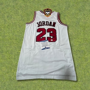 May include: White basketball jersey with the name "JORDAN" and the number "23" in red. The jersey has a red and black trim and a patch at the top. A certificate of authenticity is attached.