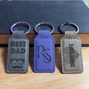 May include: Three leather keychains with different designs. The first keychain is gray and says "Best Dad" with a pair of glasses and a mustache. The second keychain is purple and has the initials "RS" with the text "Est. 2025". The third keychain is brown and has a fingerprint design with the text "Drink Coffee".