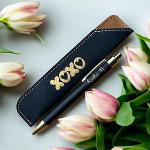 May include: A black pen and matching pen case with gold accents. The pen case is embossed with the word "XOXO" and a heart symbol. The pen is engraved with the name "Sasha N." The pen and case are displayed with pink and white tulips.
