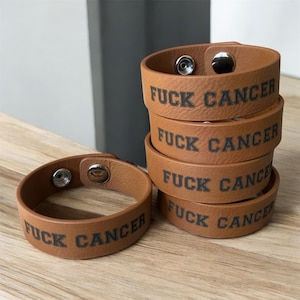 May include: A stack of brown leather bracelets with the phrase "FUCK CANCER" printed in black. Each bracelet has a snap closure. One bracelet is lying flat, while the others are stacked on a wooden surface.