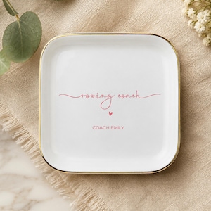 May include: A square white ceramic trinket dish with a gold-colored rim. The center features the text "rowing coach" in a pink cursive script, a small pink heart icon, and the name "COACH EMILY" printed in a simple pink sans-serif font.