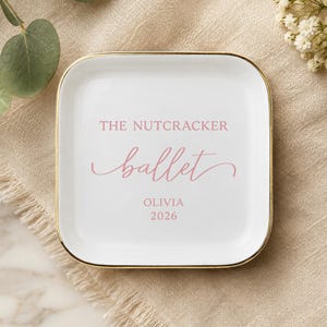 May include: A square white ceramic trinket dish with a gold-toned rim. The center features pink text that reads THE NUTCRACKER ballet, with the name OLIVIA and the year 2026 printed below in a simple sans-serif font.