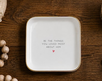 May include: A square white ceramic trinket dish with a gold-toned rim. The center of the dish features the black printed text BE THE THINGS YOU LOVED MOST ABOUT HIM with a small red heart icon positioned directly below the text.