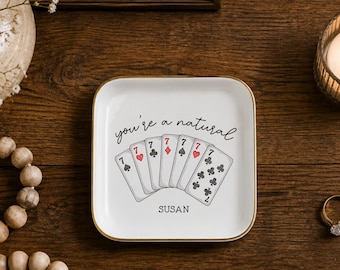 May include: A square white ceramic trinket dish with a gold-colored rim. The center features a graphic of six playing cards showing sevens, with the text "you're a natural" above the cards and the name "SUSAN" printed in black below them.