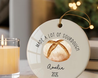 May include: A round, off-white ceramic ornament with a gold string for hanging. The ornament features a watercolor image of a sourdough loaf and the text "MADE A LOT OF SOURDOUGH", the name "Amelia", and the year "2025".