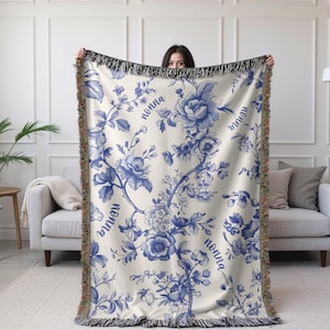 May include: Cream-colored throw blanket with a blue floral pattern and the word "nonna" repeated throughout. The blanket has a fringed edge and is held up by a person. The floral design includes roses and other flowers.