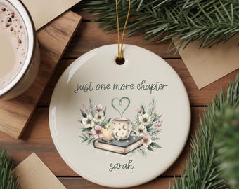 May include: A round, white ceramic ornament with a gold string for hanging. The ornament features the text "just one more chapter" and a watercolor illustration of a coffee mug, book, flowers, and a heart. The name "sarah" is written at the bottom.