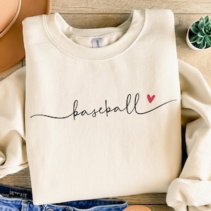 May include: A cream-coloured sweatshirt featuring the word "baseball" in a black script font and a small red heart. The sweatshirt has a crew neck and long sleeves. The image is taken from a top-down perspective.