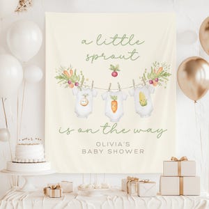 May include: A cream-colored baby shower backdrop with the text "a little sprout is on the way" in green script. The backdrop features illustrations of baby onesies with vegetable designs, and is decorated with balloons and gifts.