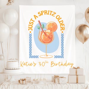 以下が含まれることがあります： 「JUST A SPRITZ OLDER」と「Katie's 30th Birthday」の文字が入った白いバナー。ストローとオレンジスライスが入ったオレンジ色のカクテルのイラストが特徴です。背景には青と金の風船があります。