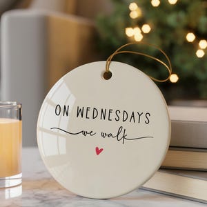 Puede incluir: Adorno redondo de cerámica blanquecina con el texto "ON WEDNESDAYS we walk" en escritura negra, un pequeño corazón rojo y una cuerda dorada para colgar. El adorno está delante de una pila de libros y una vela encendida.