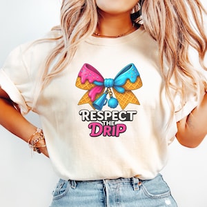 May include: Cream-colored t-shirt featuring a colorful graphic of an ice cream cone bow with pink and blue ice cream, and the text "Respect the Drip". The design includes a blue megaphone and a blue pom-pom.