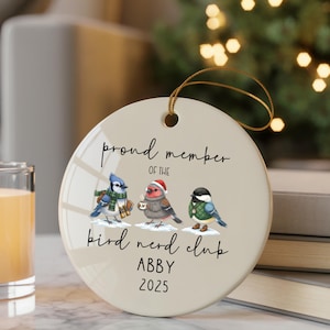 May include: A round, cream-colored ceramic ornament with a gold string for hanging. The ornament features three illustrated birds and the text "proud member of the bird nerd club ABBY 2025."