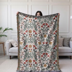 May include: A woven throw blanket featuring a repeating pattern of red flowers, mushrooms, and suns on a cream background. The design includes green leaves and stems. The blanket has a fringed edge.