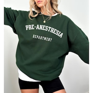 May include: A dark green sweatshirt with the words "PRE-ANESTHESIA DEPARTMENT" printed in white block letters. The sweatshirt has a crew neck and long sleeves. The person is wearing a white undershirt and black shorts.