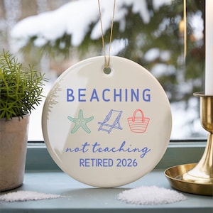 May include: A round, off-white ceramic ornament with the words "BEACHING not teaching RETIRED 2026" in blue script. The ornament features a starfish, beach chair, and tote bag graphic. A gold string is attached for hanging.
