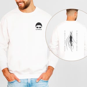 Peut inclure: Sweat-shirt blanc avec une silhouette noire de nageur au dos et le texte "ALWAYS LOOKING UP". Le devant du sweat-shirt présente une silhouette noire de nageur portant un bonnet de bain et des lunettes de natation avec le texte "CHARLIE" en dessous.
