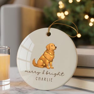 May include: A round ceramic Christmas ornament with a golden retriever dog wearing fairy lights. The ornament has the text "merry & bright" and the name "Charlie". A gold string is attached for hanging.