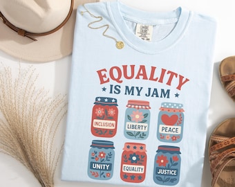 May include: Light blue t-shirt with the text "EQUALITY IS MY JAM" and illustrations of six jars, each with a different word: Inclusion, Liberty, Peace, Unity, Equality, and Justice. A gold necklace and a beige hat are also in the image.