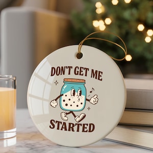 May include: A round, cream-colored ceramic ornament with the phrase "DON'T GET ME STARTED" and a cartoon jar of milk with arms and legs. The jar is light blue with a brown lid and is walking with a musical note. A gold string is attached for hanging.
