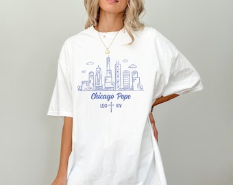 Könnte beinhalten: Weißes T-Shirt mit blauem Skyline-Grafikdesign, dem Text "Chicago Pope" und "LEO XIV". Das Design beinhaltet ein Kreuz. Das Shirt hat eine lockere, übergroße Passform.