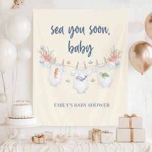 May include: A cream-colored baby shower banner with the text "sea you soon, baby" in navy blue script. Three onesies with sea creature illustrations hang from a clothesline. The banner also includes the text "EMILY'S BABY SHOWER".