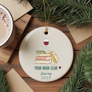 May include: A round, white ceramic ornament with a gold string for hanging. The ornament features a stack of books, a wine glass, and the text "YOUR BOOK CLUB" with a heart, the name "Ashley", and the year "2025".