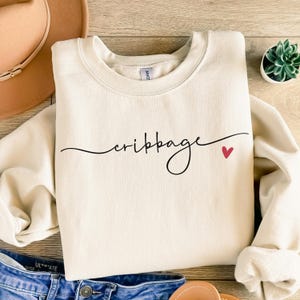May include: A cream-colored crewneck sweatshirt featuring the word cribbage written in black cursive script across the chest, accompanied by a small red heart icon positioned to the right of the text.