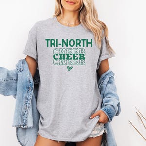 May include: A light gray t-shirt with the words "TRI-NORTH CHEER CHEER CHEER" in green lettering. The shirt has a relaxed fit and short sleeves. A small green heart is below the text.