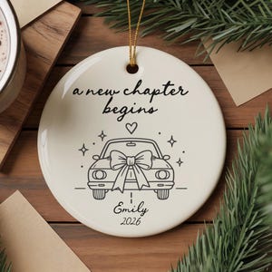 May include: A round white ceramic ornament featuring a black line drawing of a car with a large bow on the hood. The text reads a new chapter begins, Emily, and 2026. A small heart and star icons are included. It hangs from a gold string.