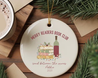 May include: A round, white ceramic ornament with a gold string for hanging. The ornament features the text "MERRY READERS BOOK CLUB" and "read between the wines". A stack of books, a wine bottle, and the name "Addie" are also displayed.