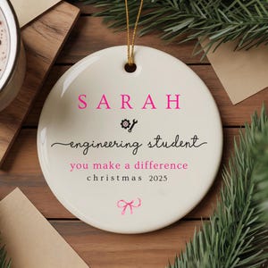 May include: A circular, cream-coloured ceramic Christmas ornament with the name "SARAH" in pink, and the words "engineering student" and "you make a difference christmas 2025". A gold string is attached. The ornament features a gear icon and a pink bow.