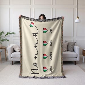 May include: Cream-colored woven throw blanket with black fringe and the word "Nonna" in a script font. The blanket features heart-shaped designs with the colors of the Italian flag and names: Kasey, Rose, Charlie, and Olivia.