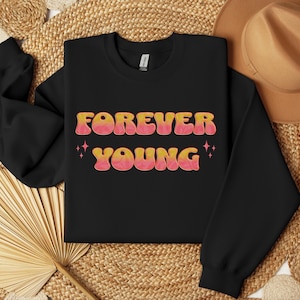 Op de afbeelding: Zwart sweatshirt met de tekst "FOREVER YOUNG" in roze en oranje retro letters met glinsterende sterren aan beide kanten.