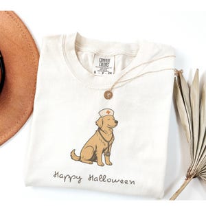 Puede incluir: Camiseta color crema con un golden retriever de dibujos animados con gorro de enfermera y estetoscopio. La frase "Happy Halloween" está escrita debajo. También se ven un collar dorado y un sombrero marrón. La camiseta es de algodón.